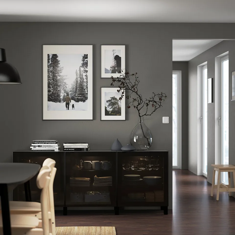 Modern dining room with dark grey walls, GLASSVIK cabinet, prints, chair, vase with twigs, and natural light.