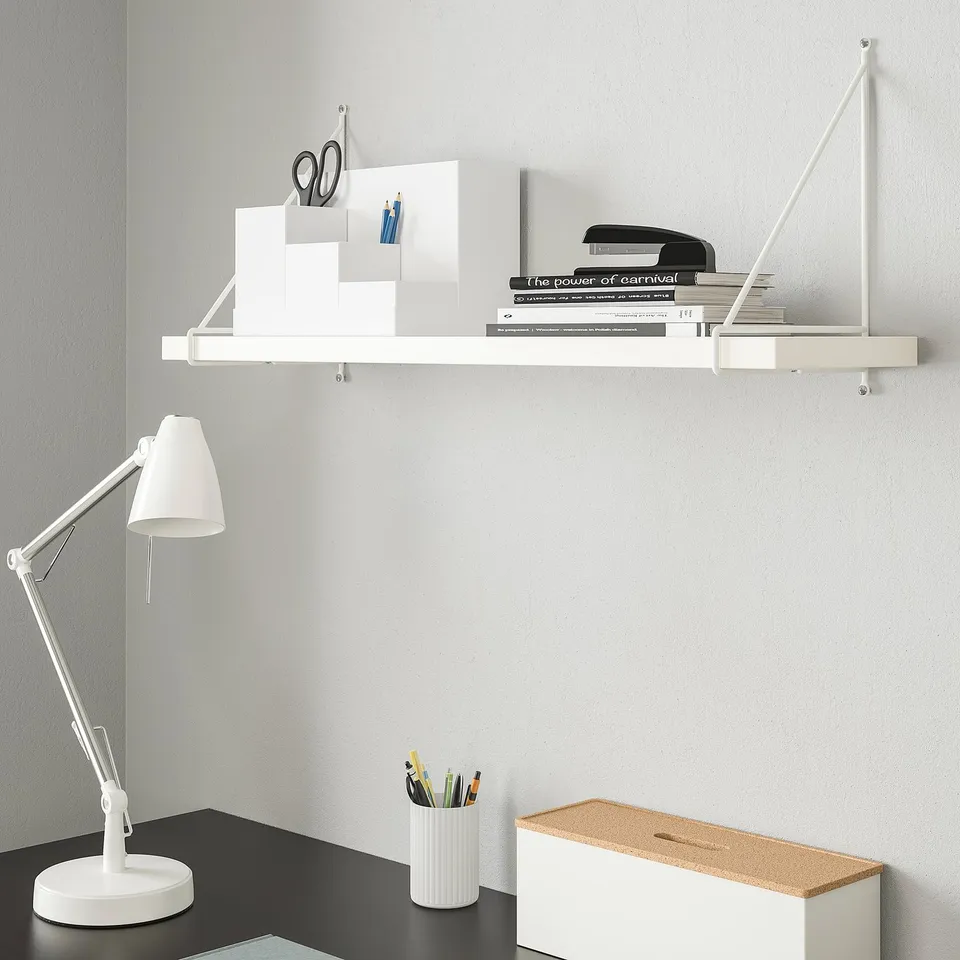 BERGSHULT shelf with office supplies on a light grey wall, above a black desk with a lamp and storage box.
