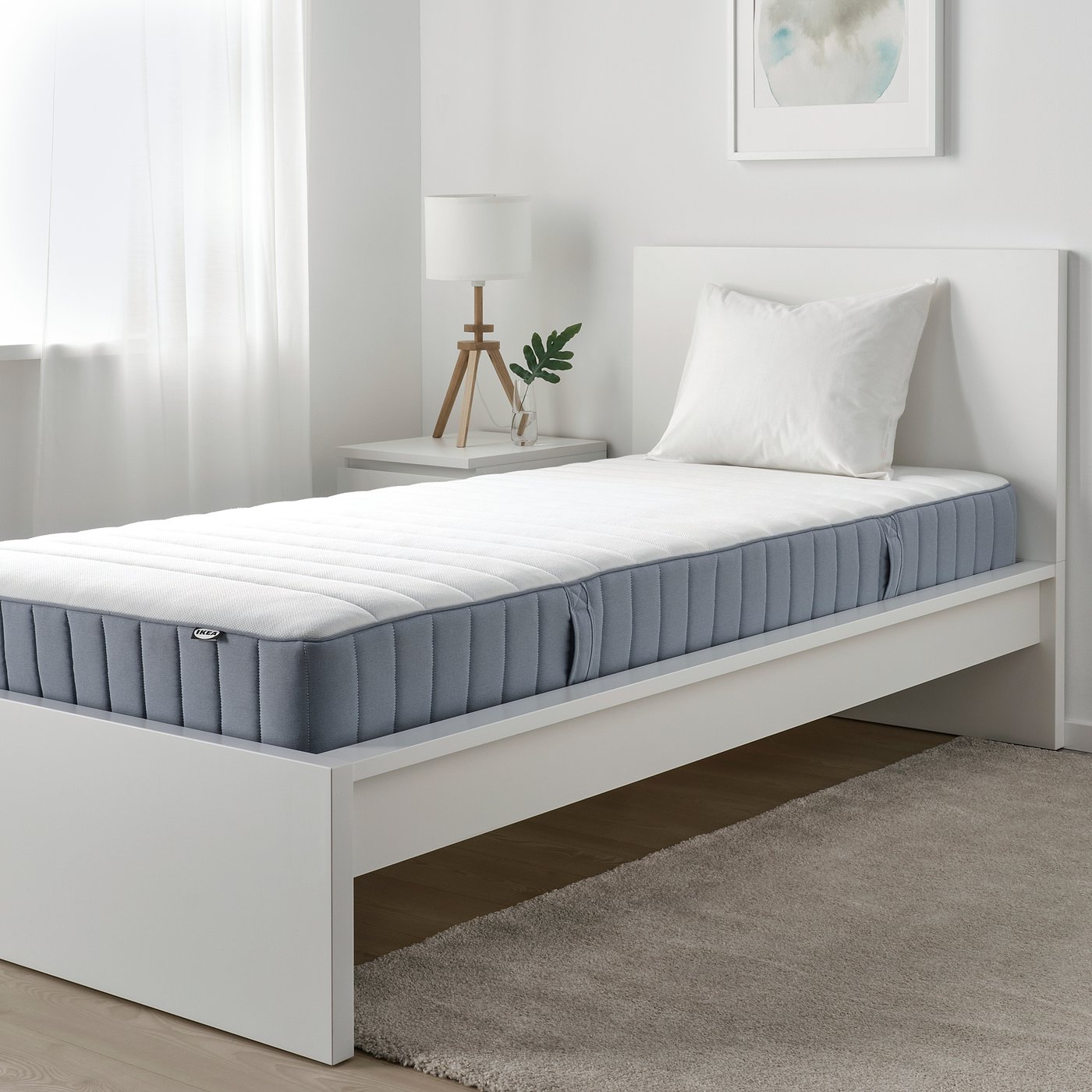 Modern minimalist bedroom. Single white bed with grey mattress, white pillow, stylish lamp, small plant, grey rug. Wall art. Soft, neutral tones.