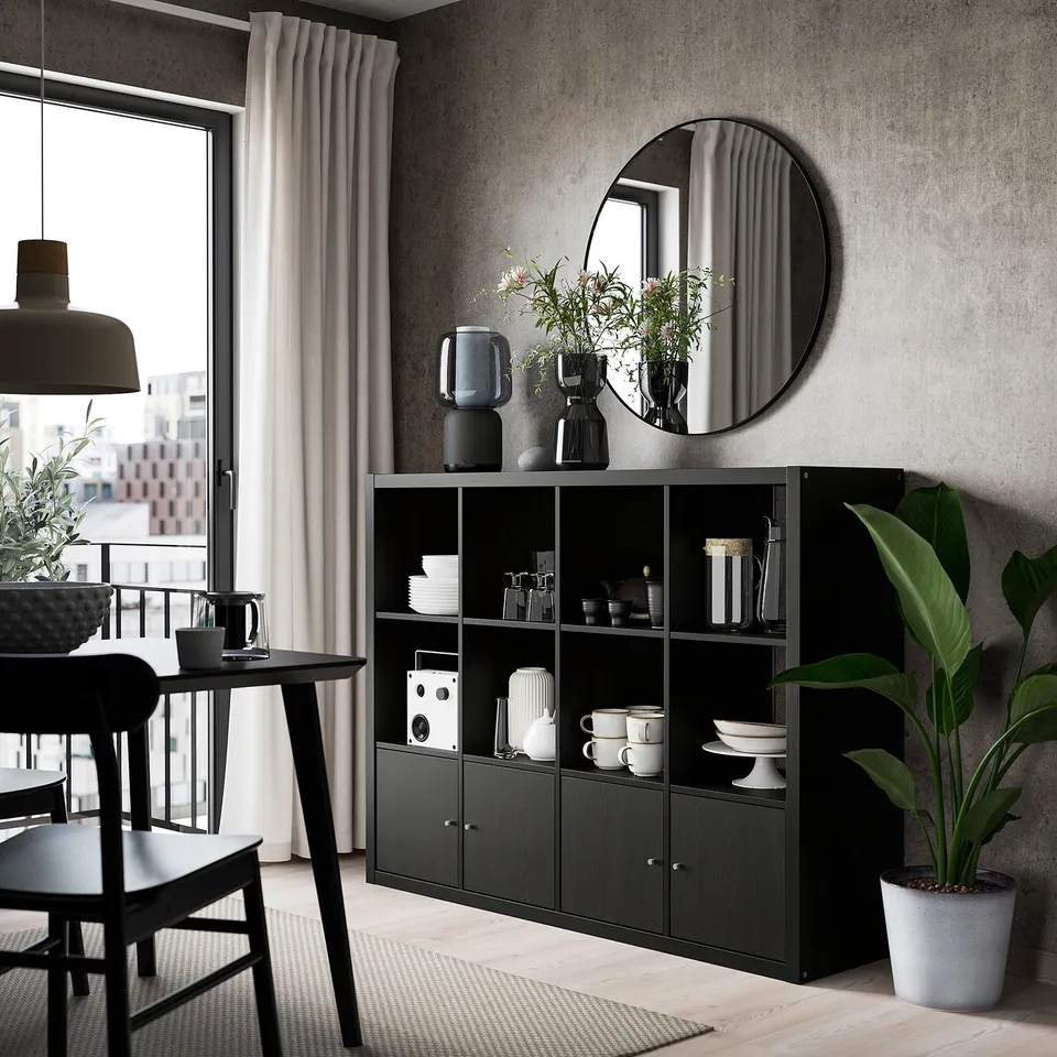 Black KALLAX shelf unit holds dishes and plants, positioned beside a window, between curtains and a chair.