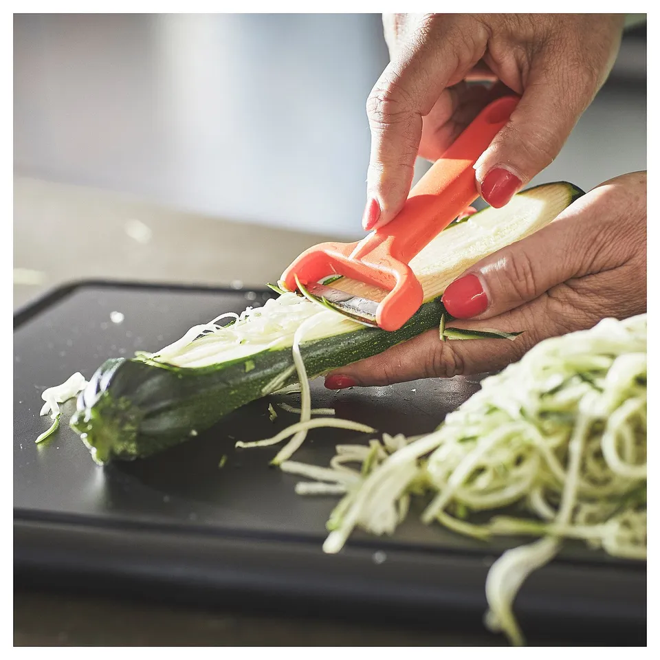 Person peels courgette with bright orange UPPFYLLD peeler. Cucumber strips on black board, nails painted red.