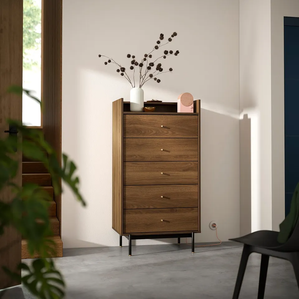 Wooden chest with five drawers, sleek brass handles. Decorated with vase and flowers. Positioned near window for natural light.