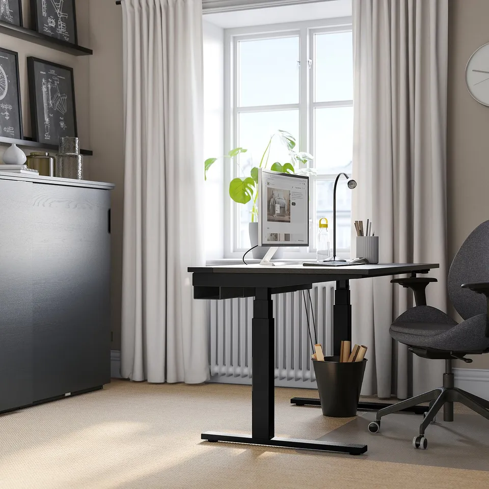 Modern home office with MITTZON black sit-stand desk, grey chair, large windows, white curtains, and grey carpet. Desk holds pc, lamp, and plant.
