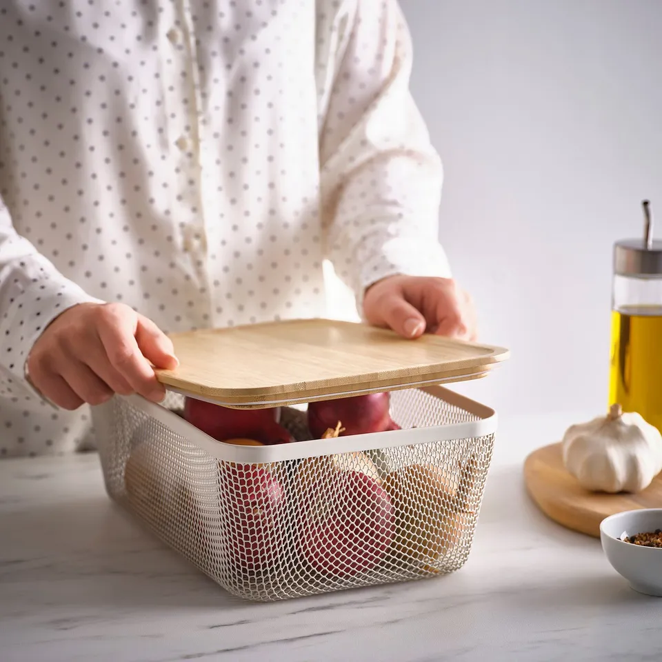 Person opens mesh basket with lid for food storage, displaying fresh produce. IKEA 365+ storage solution allowing air circulation and easy visibility.