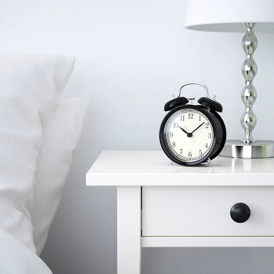 Black alarm clock on white bedside table, next to white lamp and bed with white pillow. Silent quartz movement.