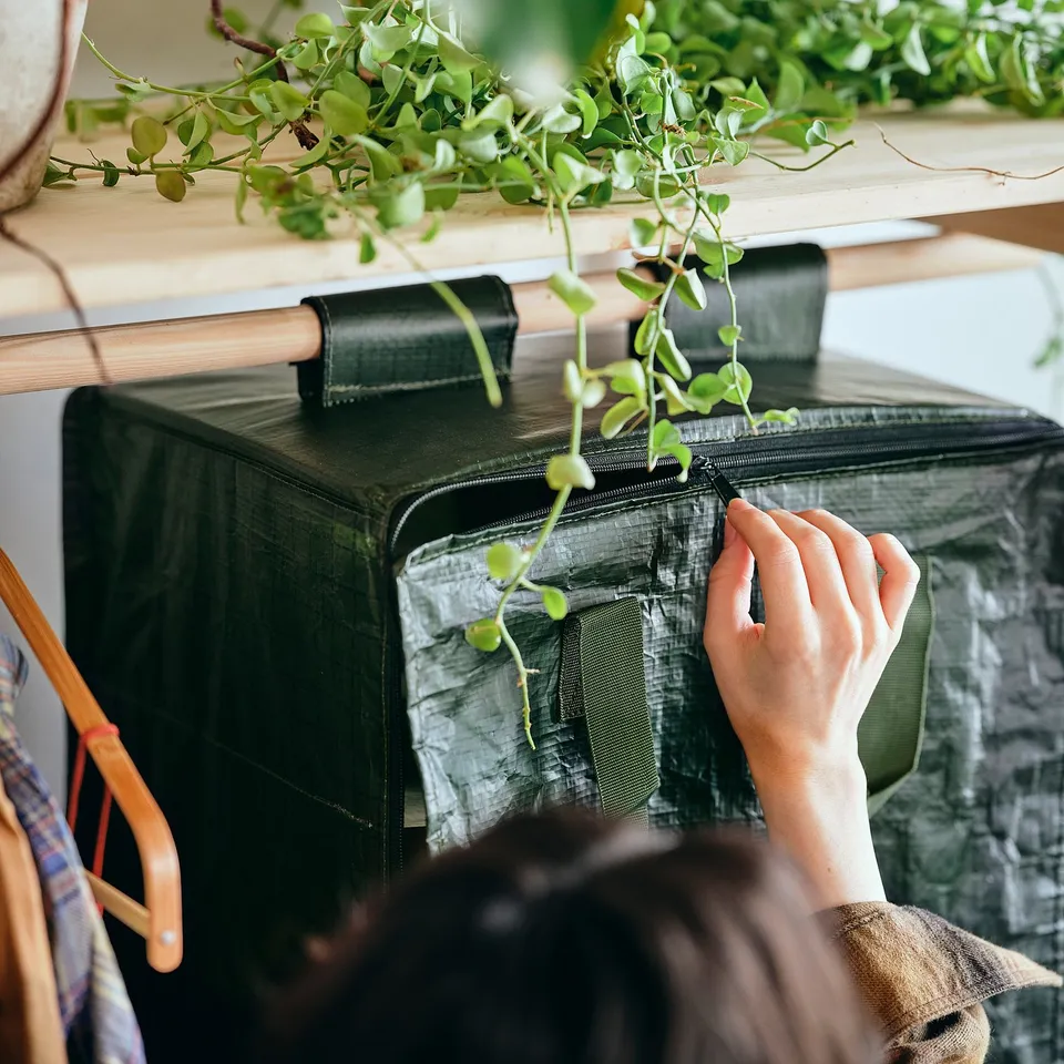 A person is opening a large hanging storage bag made of fabric, with visible stitching and straps, placed under a shelf with trailing green plants.