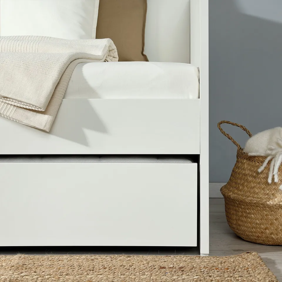 White daybed with trundle drawer, soft pillows, and woven basket for storage.