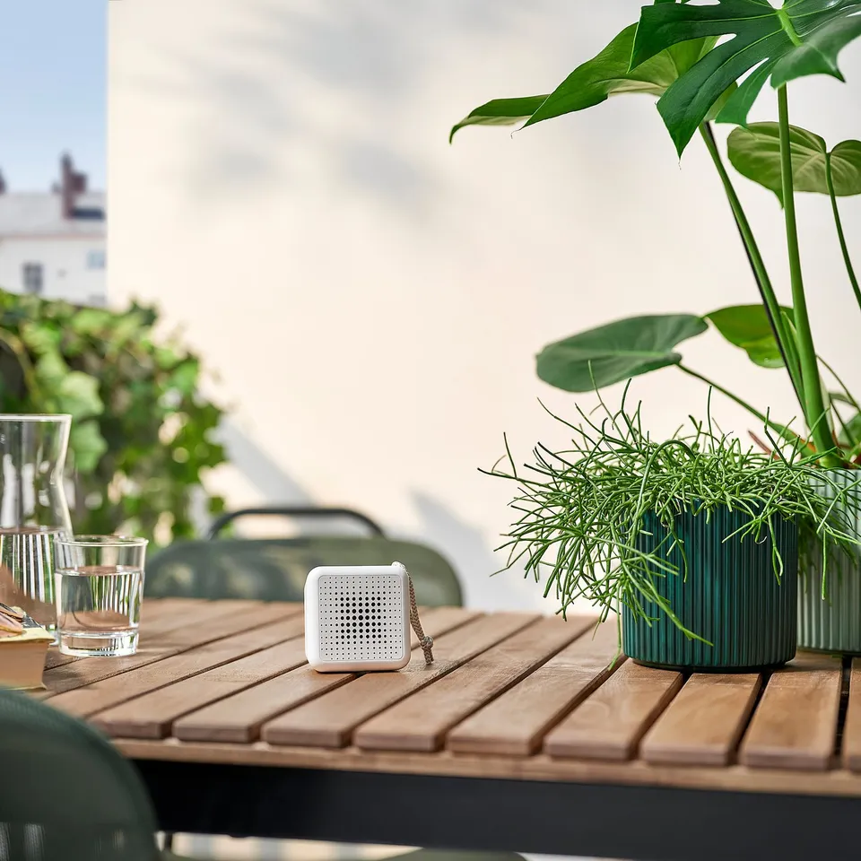 Small white VAPPEBY speaker on wooden table with green plants. Lightweight, portable design perfect for outdoor use.