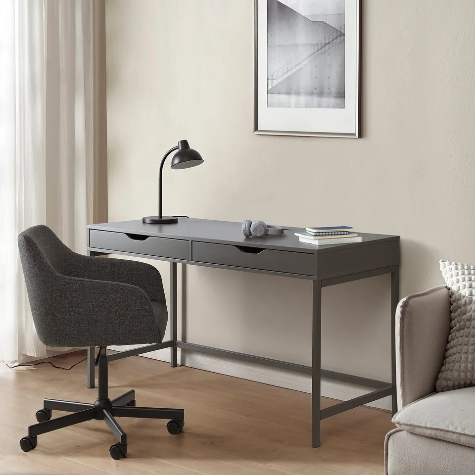 Small black ALEX desk with drawers in a room, next to a grey chair. A lamp, headphones, and books on top. Its compact and sleek, designed for storage.