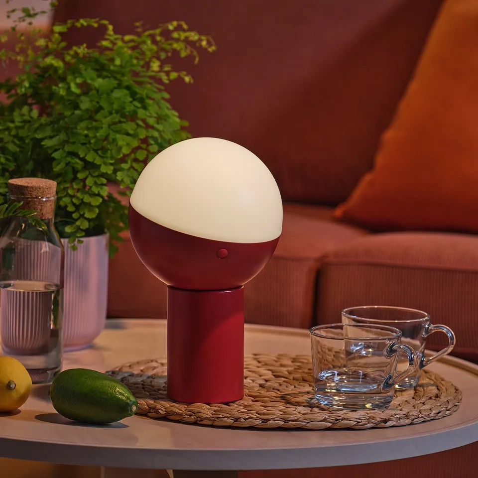 Modern table lamp with half-globe shade. Brightening cosy space with a red base. Surrounded by cups and fruits on a side table.