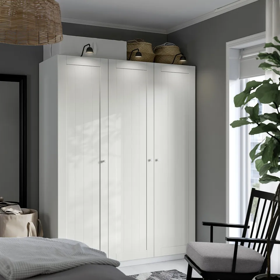 A white PAX wardrobe with sleek, modern cabinets and minimalist design. It stands tall, occupying much of the space, with woven baskets atop.