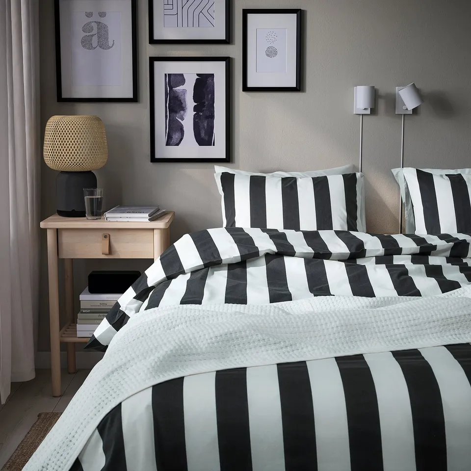 Bed with black/white striped SLÅNHÖSTMAL duvet, art wall, bedside table, lamp, books, and glass.