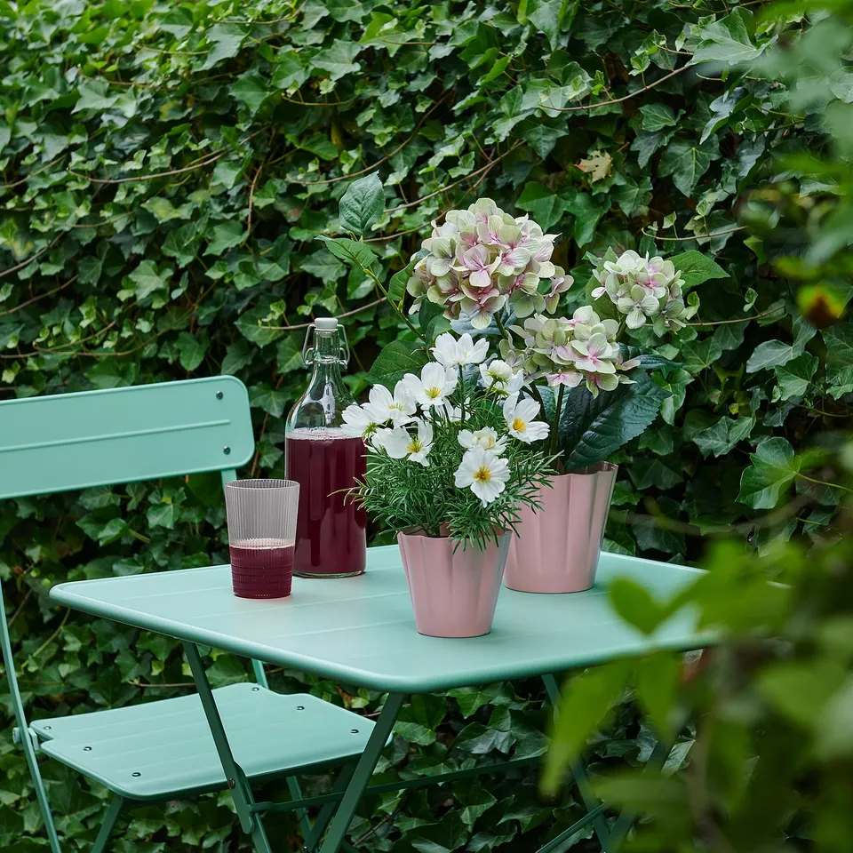 Green table & chairs with pink plants, flowers, & glassware against greenery.