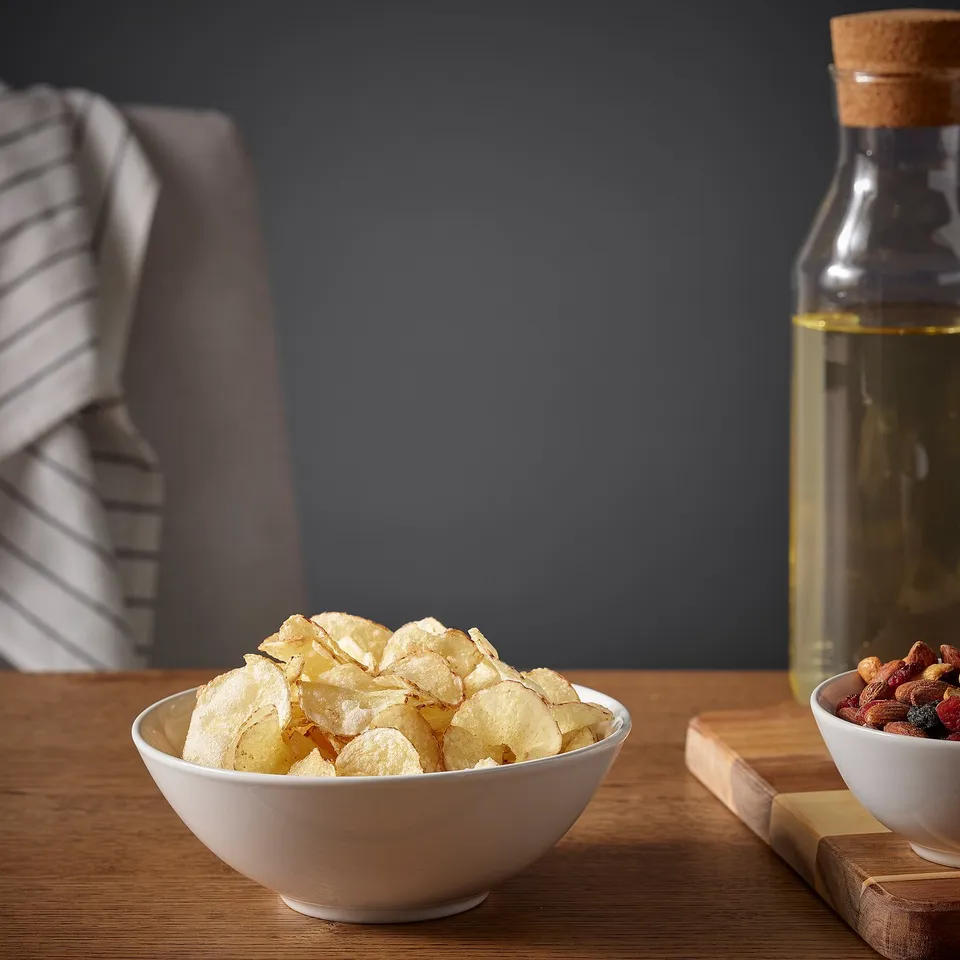 Bowl of potato chips on wooden table with oil bottle and mixed nuts; dining setup.