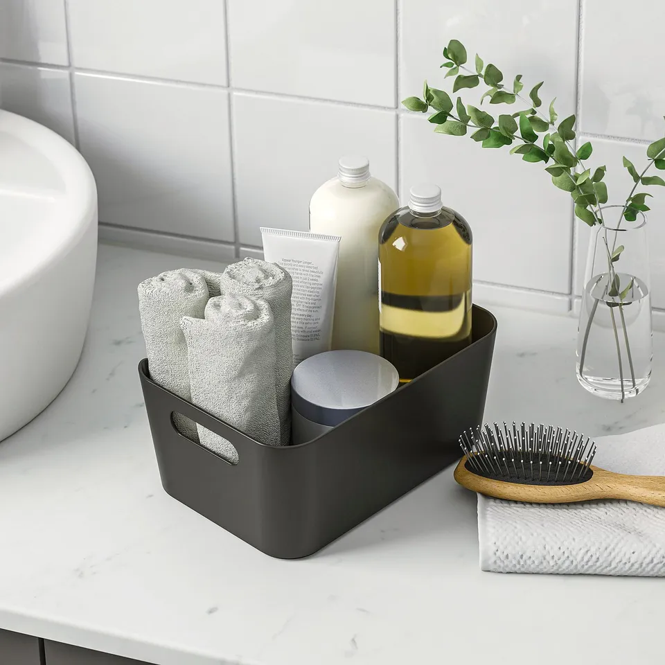 Dark plastic storage bin organises toiletries. Contains rolled towels and bottles on white marble counter, with handles for easy carrying.