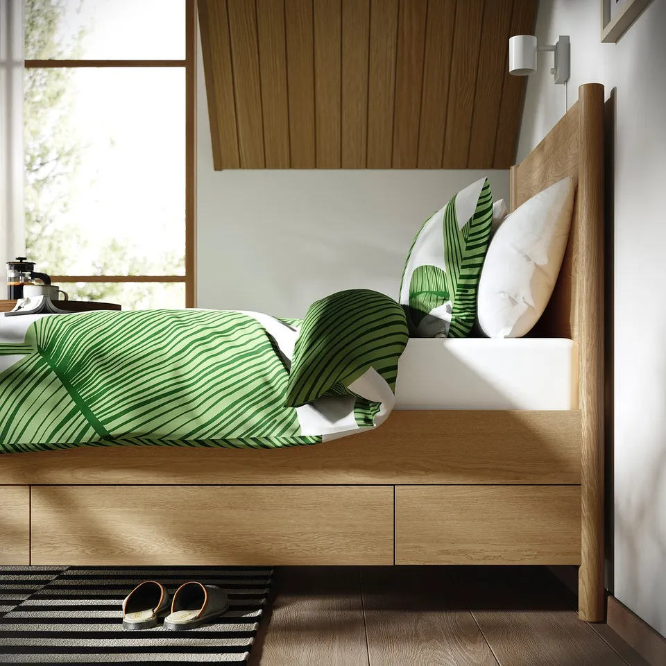 A modern wooden bed with soft-close drawers beneath, featuring a green striped comforter and tropical leaf pillows, set against a wooden panel wall.