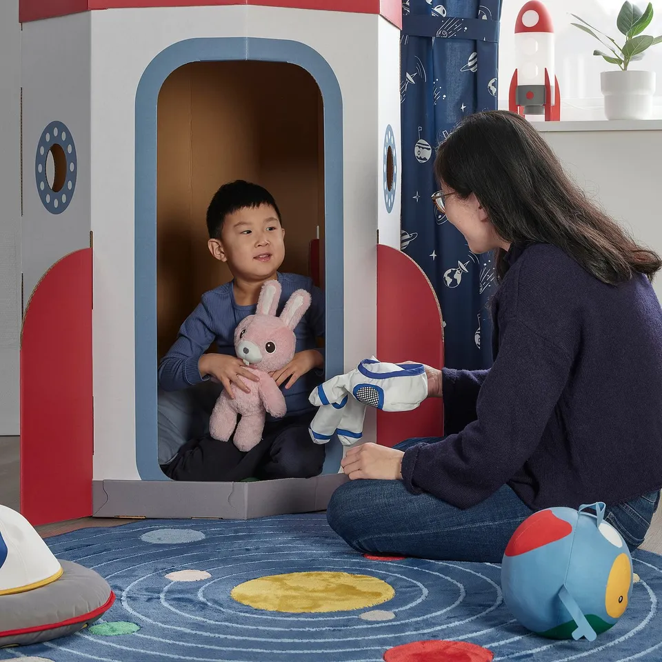 Person & child play in cardboard space station, toys in hand.