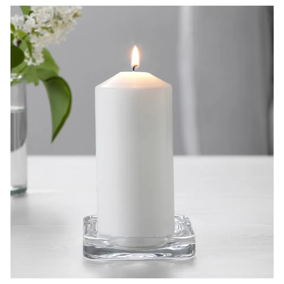 A lit white candle sits in a clear square glass holder, creating ambiance on a wooden table.