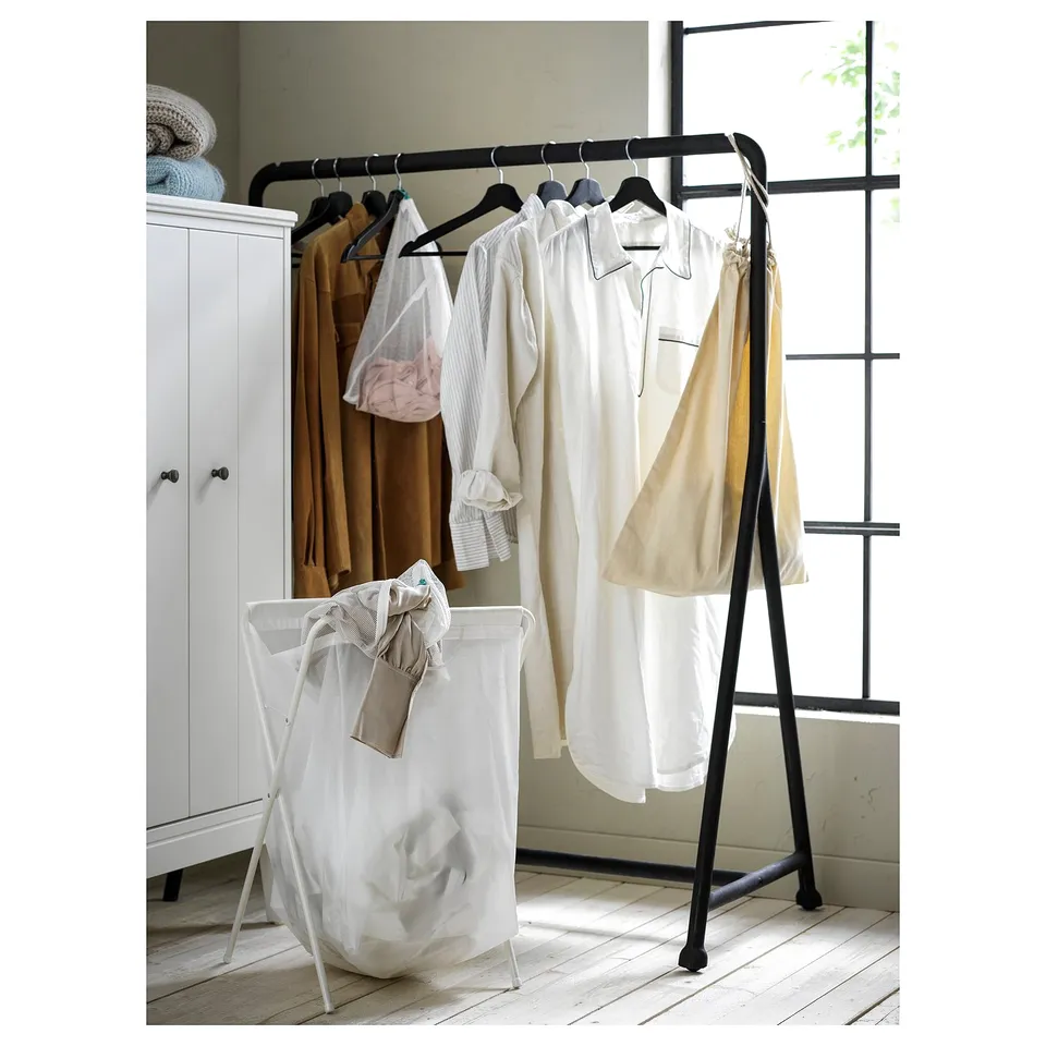 Black metal clothing rack with wheels, holding light-colored clothes near a white cabinet and a white laundry basket.