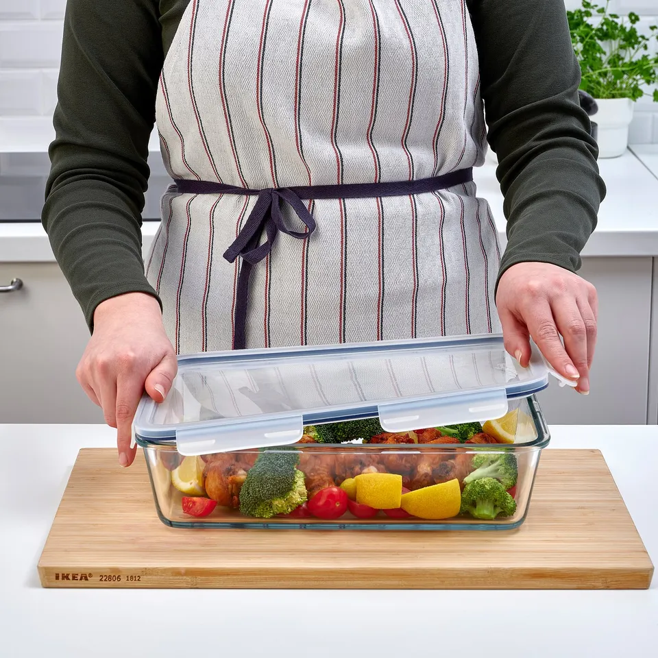 Person placing lid on IKEA 365+ glass food container filled with roasted chicken and vegetables on a bamboo cutting board.