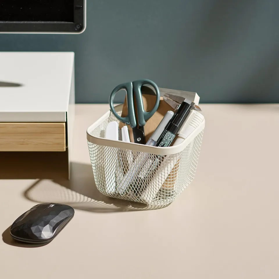 A white mesh office organiser holds scissors, markers, and a ruler on a desk. A black mouse is beside it.