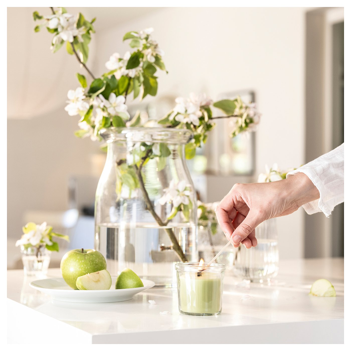 Green candle lit next to vase with white flowers, apples on marble table.
