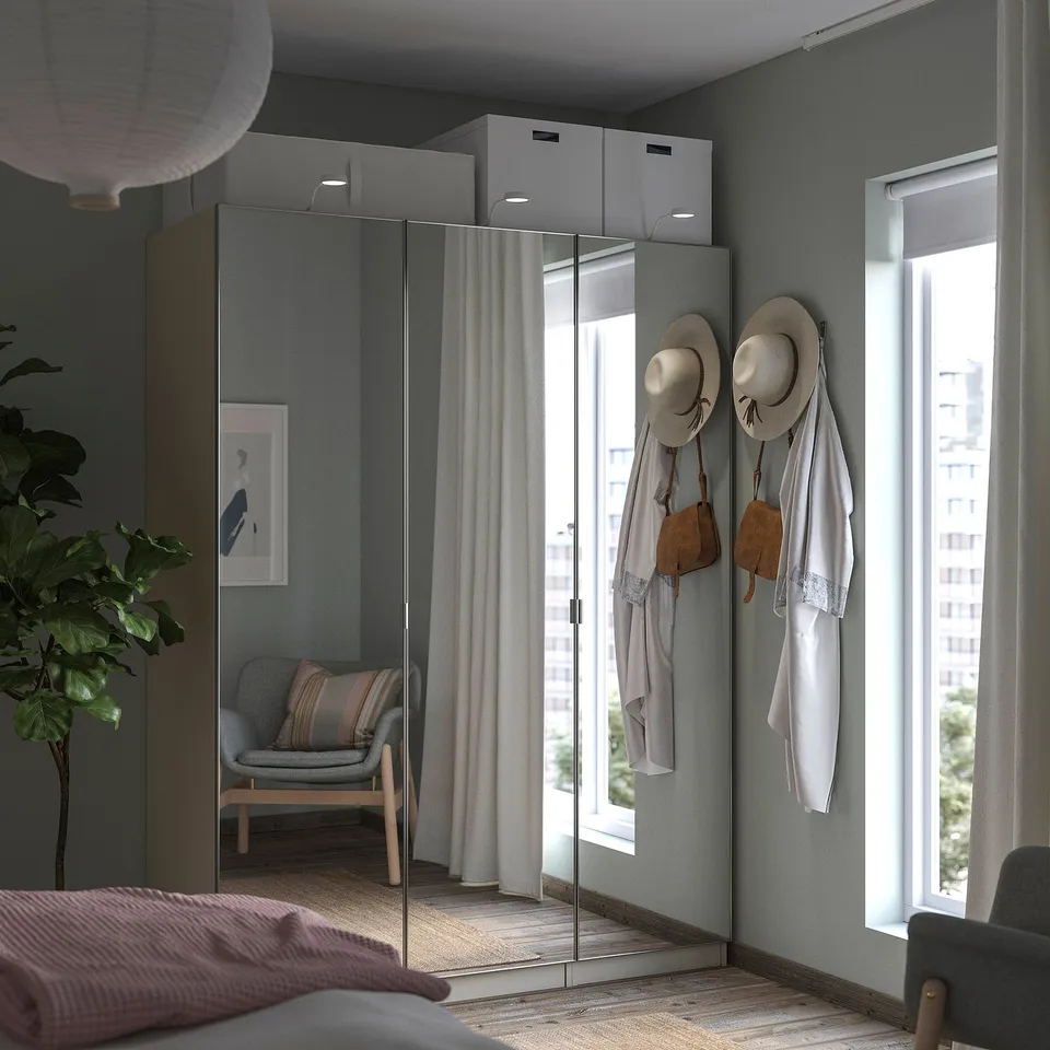 Modern bedroom a sleek grey PAX wardrobe with mirrored doors, white curtains, and potted plants, offering a minimalist and organised space.