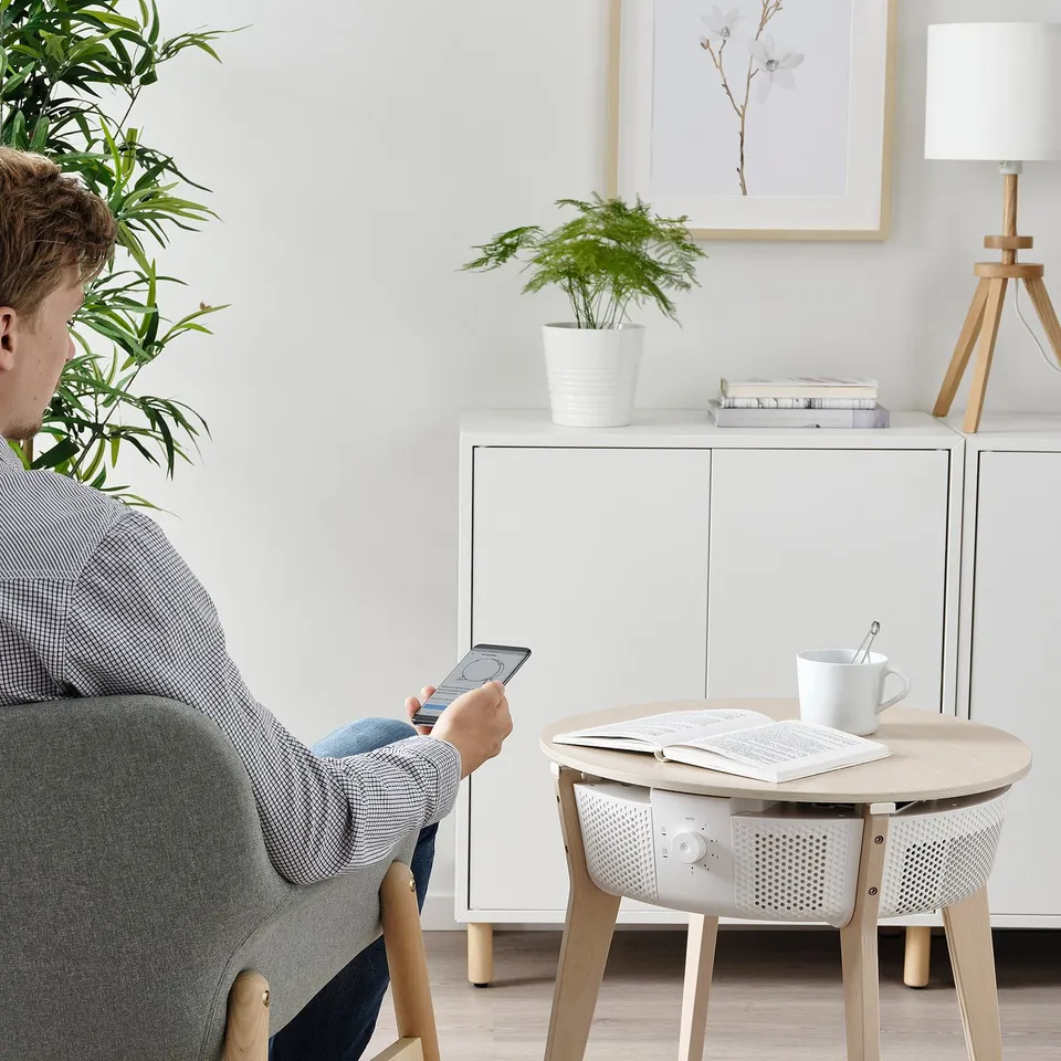 Person sitting on chair using smartphone, next to a table with air purifier, cup, and book.
