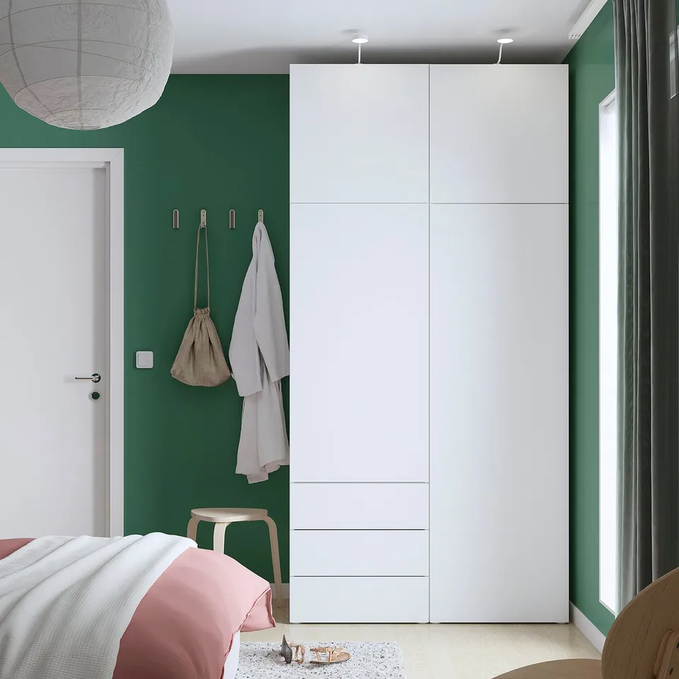 White PLATSA wardrobe in green room. Tall, sleek, with multiple compartments and towels. Near bed, stool, and large window.