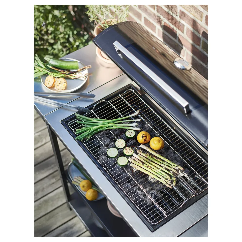 Grilling veggies on GRILLSKÄR barbecue. Stainless steel grill with asparagus, courgette, lemons. Black bbq on wood deck with brick wall.