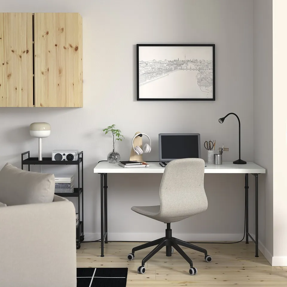 Modern home office with white desk, black legs, & grey chair. Computer, lamp, & accessories on desk. Minimalist art print & bookshelf.