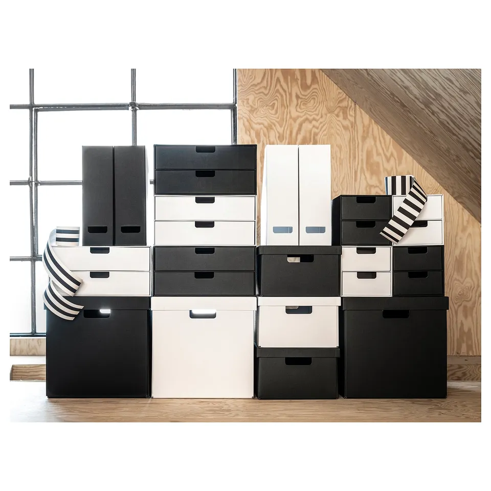 Stacked black and white storage boxes. Boxes vary in size, arranged neatly on wooden flooring and plywood wall. Modern and minimalist design.