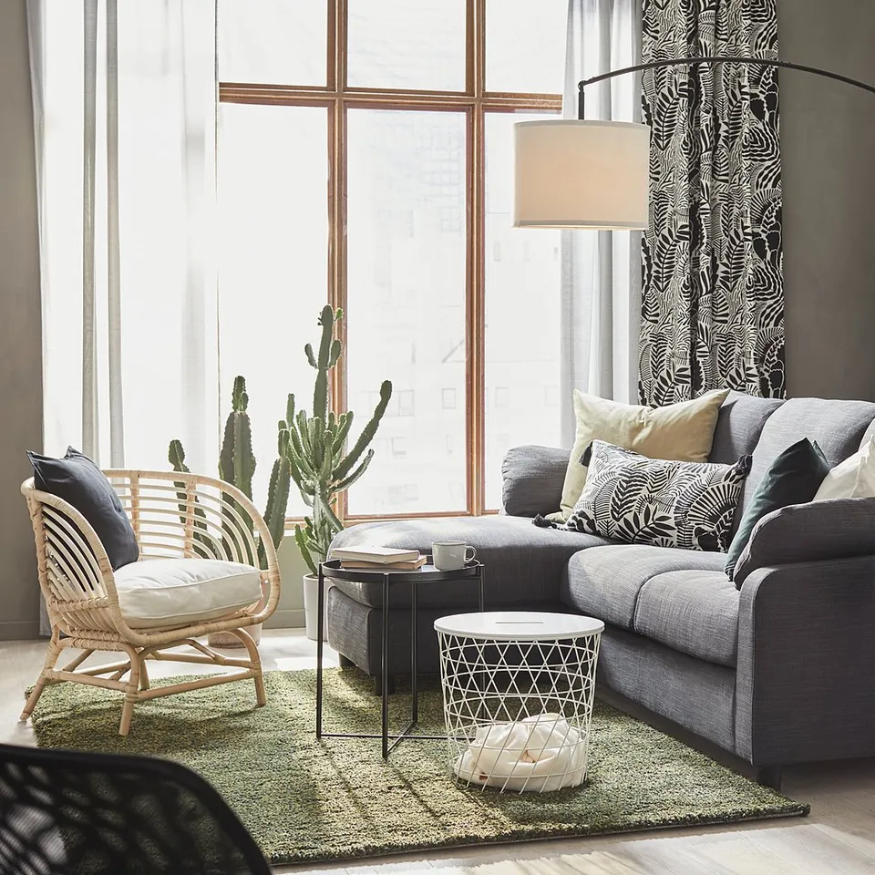 Modern living room with grey SKAFTET floor lamp, wicker chair, grey sofa with pillows, glass tables, and large window with curtain.