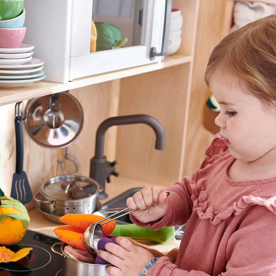 Girl in pink sweater plays with toy kitchen, cooking colourful veggies.