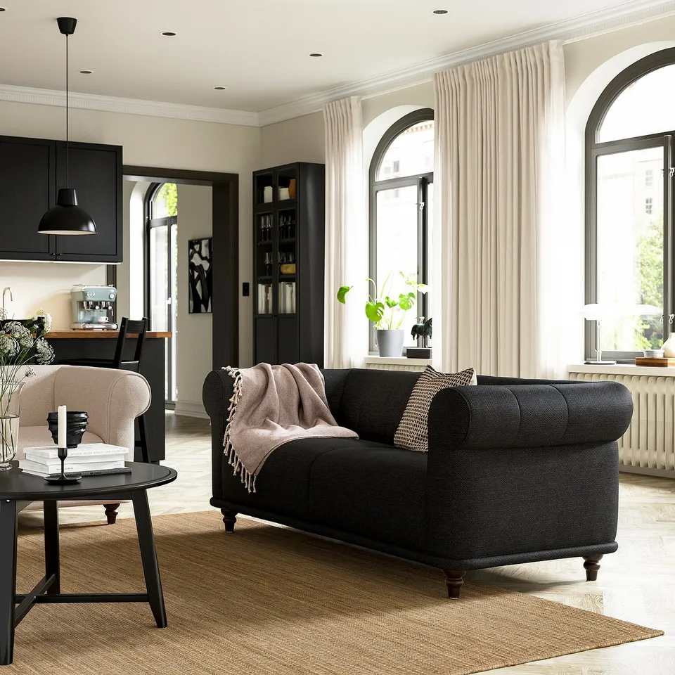Elegant living room with black VISKAFORS sofa, soft cushions, large windows, and modern decor. Light streams in, highlighting the cosy ambiance.