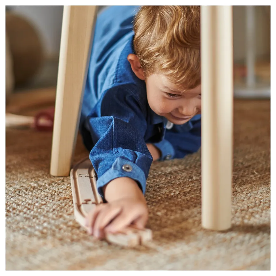 Young child playing under wooden chair, wearing blue jeans and holding wooden toy.