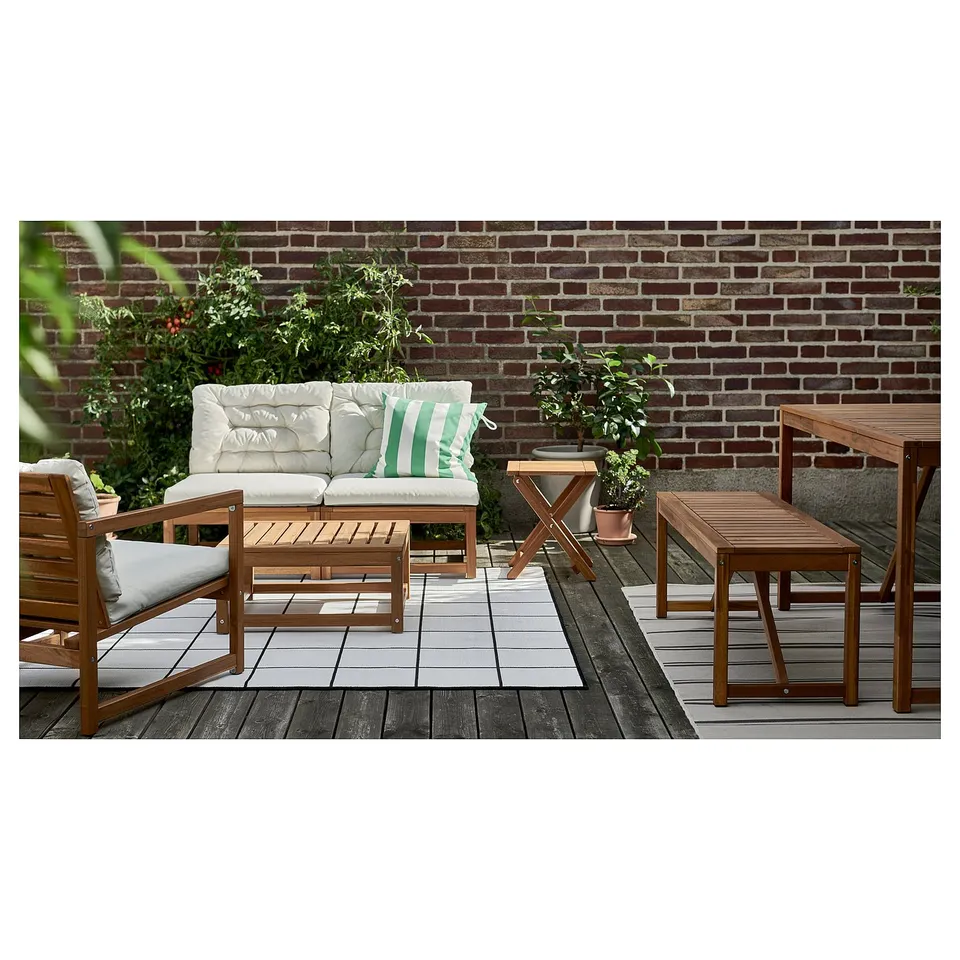 Outdoor patio with NÄMMARÖ wooden furniture: sofa with beige cushions, coffee table, bench, chairs, and decorative plants, against a brick wall.