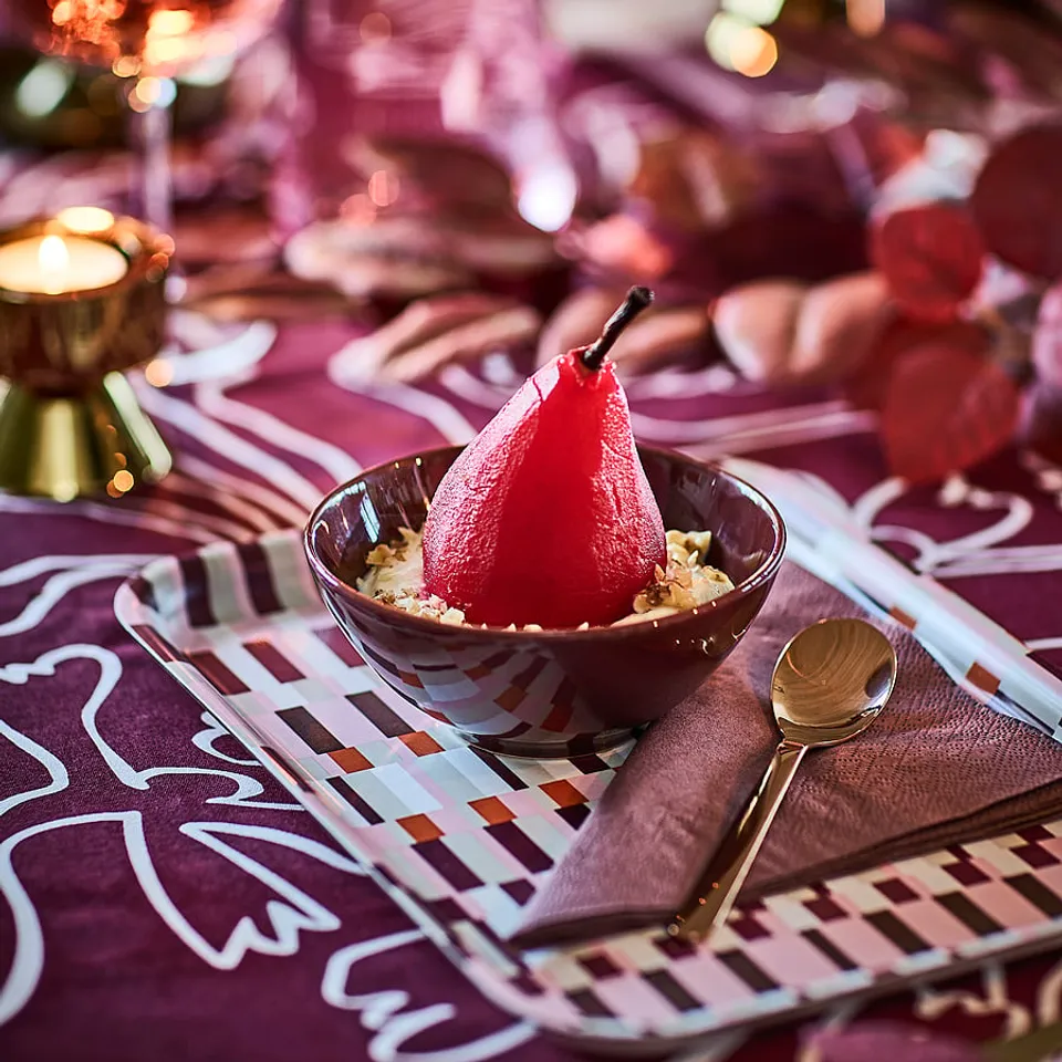 Bright red poached pear in dark bowl atop patterned purple placemat with metal cutlery, part of stylish table setting.