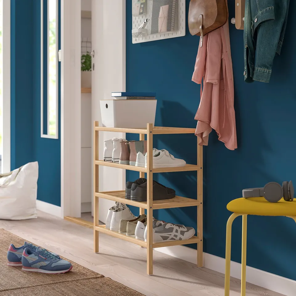 Entryway with light wooden shoe rack holding various pairs of shoes. Dark blue wall backdrop with hanging coats, a yellow stool, and scattered shoes.