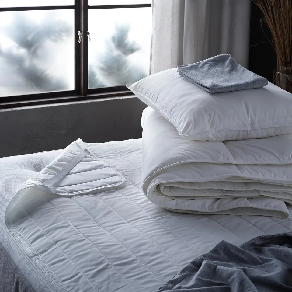 Displays a neatly arranged bed with white pillows and a folded white blanket, placed near a window with curtains.
