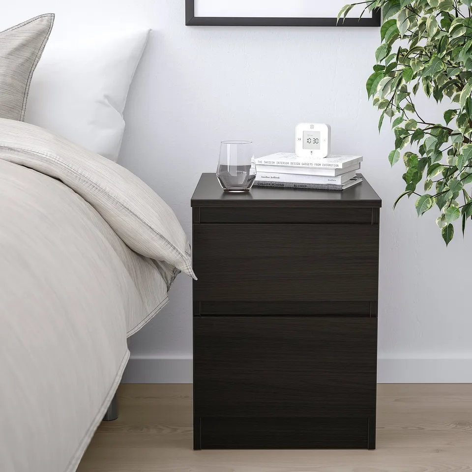 A KULLEN black bedside table with drawers, a glass, books, and clock. Minimalist design beside a bed with plant and neutral bedding.