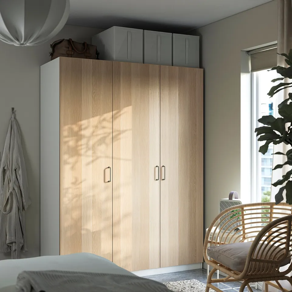 Minimalist bedroom with large light wood PAX wardrobe, grey top cabinets, and wicker chair by window.