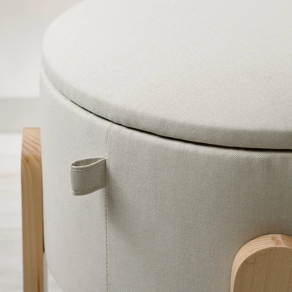 Round, upholstered stool with smooth, durable fabric. Small handle for moving, set on light wood base.