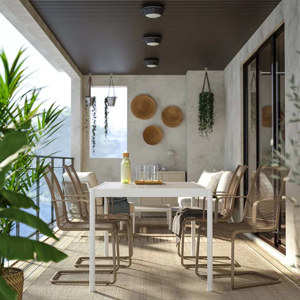 Modern patio with white table & VÄSMAN brown wicker chairs, grey cushions. Decorated with hanging plants, wall art, & outdoor rug.