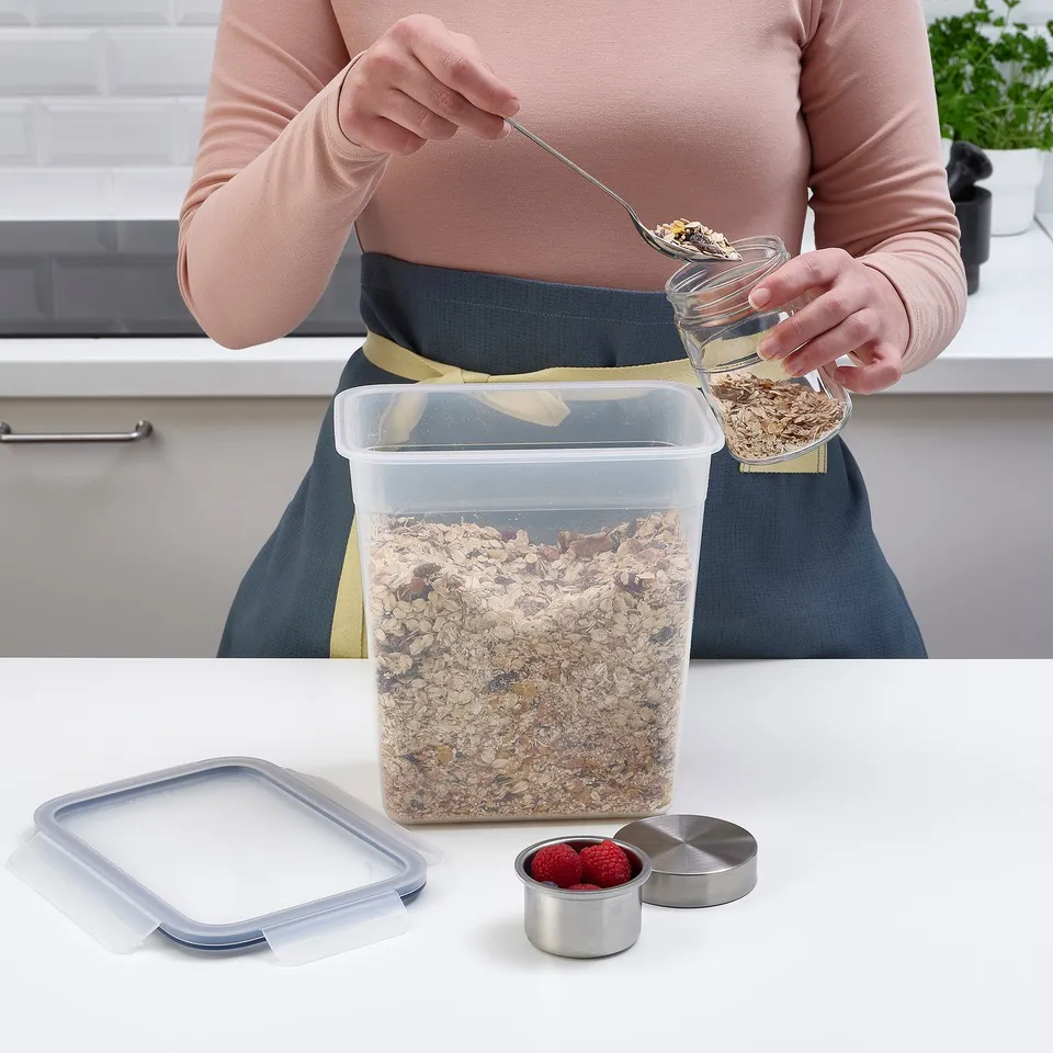 A person scoops granola from a large, clear plastic container on a kitchen counter, next to a small container of raspberries.