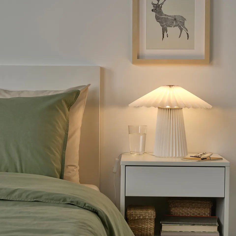 A cosy bedroom with a lit KUDDLAVA lamp on a bedside table, framed deer art, and green bedding.
