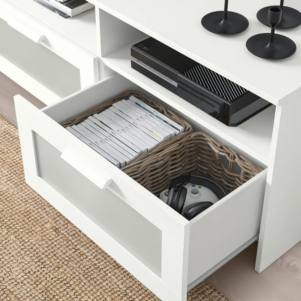 White tv stand with drawers. One drawer open showing dvds and a game controller in a basket. Electronics and candles on top.