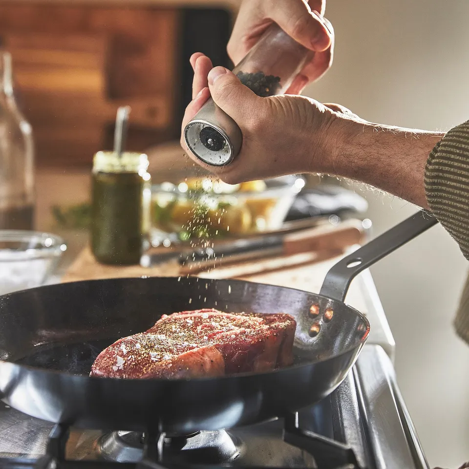 A person is seasoning a steak in a carbon steel pan on a stovetop.