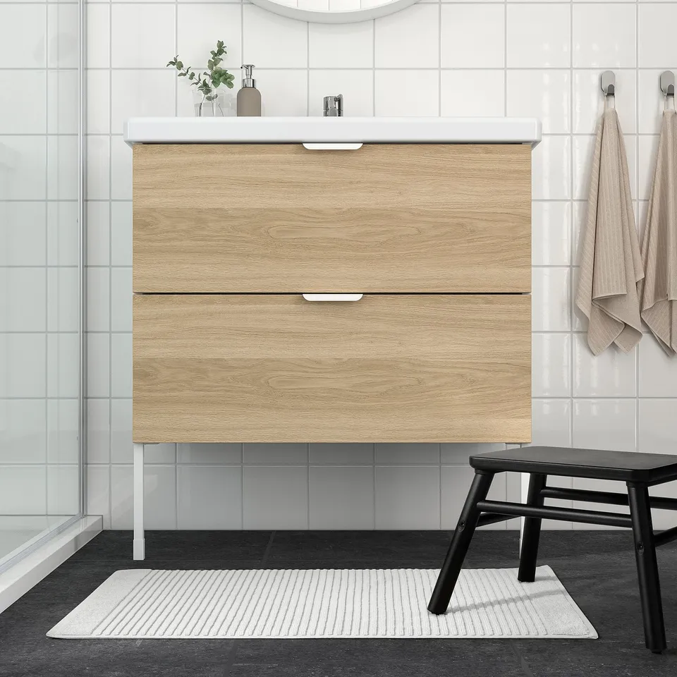 Modern bathroom with light wood vanity, white mat, black stool.