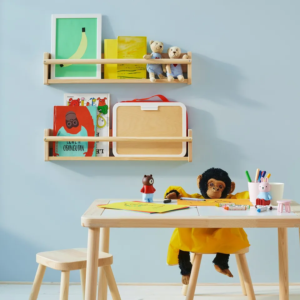 Childs play area with wall shelves holding books and stuffed toys, a table with art supplies and a monkey cuddly toy seated at it.